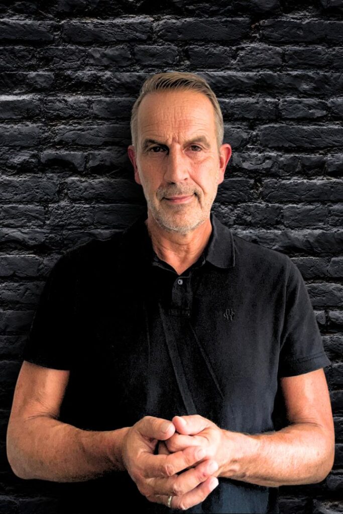 Mentalcoach Arnd Hebestreit steht vor einer Wand und faltet die Hände. Er bietet Business Coaching gegen Stress und Mentalcoaching an.