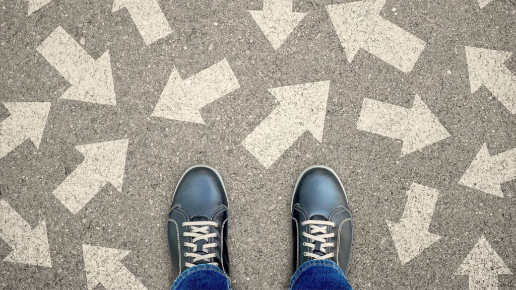 Beine einer Person in schwarzen Schuhen auf grauem Asphalt, umgeben von unzähligen in alle Richtungen weisenden Pfeilen, als visuelles Symbol für die blockierte Entscheidungsfindung unter Stress.