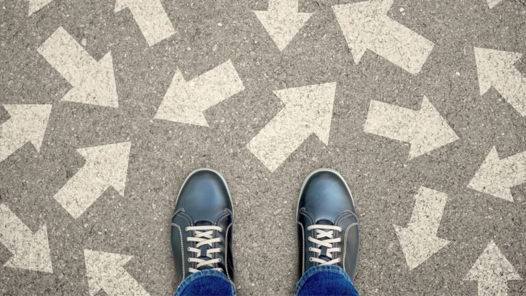 Beine einer Person in schwarzen Schuhen auf grauem Asphalt, umgeben von unzähligen in alle Richtungen weisenden Pfeilen, als visuelles Symbol für die blockierte Entscheidungsfindung unter Stress.
