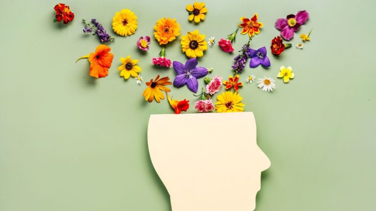 Eine Papiersilhouette eines menschlichen Profils auf grünem Hintergrund, aus dessen geöffnetem Kopf unzählige bunte Blumen sprießen. Das Bild visualisiert die aufblühende geistige und körperliche Gesundheit durch ein professionelles Mentalcoaching im Business.