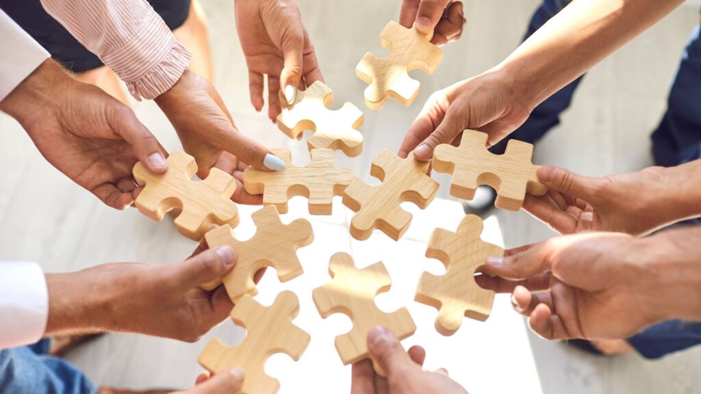 Mehrere Hände von Geschäftsleuten fügen in der Mitte eines Kreises passgenaue helle Holz-Puzzleteile zusammen. Die Grafik symbolisiert, wie ein fundiertes Resilienztraining Unternehmen stärkt, indem es isolierte Manager zu einem kollektiv belastbaren Team vereint.