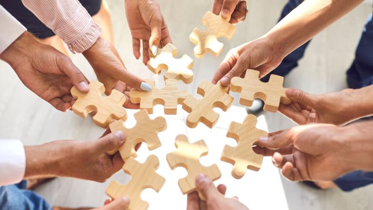 Mehrere Hände von Geschäftsleuten fügen in der Mitte eines Kreises passgenaue helle Holz-Puzzleteile zusammen. Die Grafik symbolisiert, wie ein fundiertes Resilienztraining Unternehmen stärkt, indem es isolierte Manager zu einem kollektiv belastbaren Team vereint.